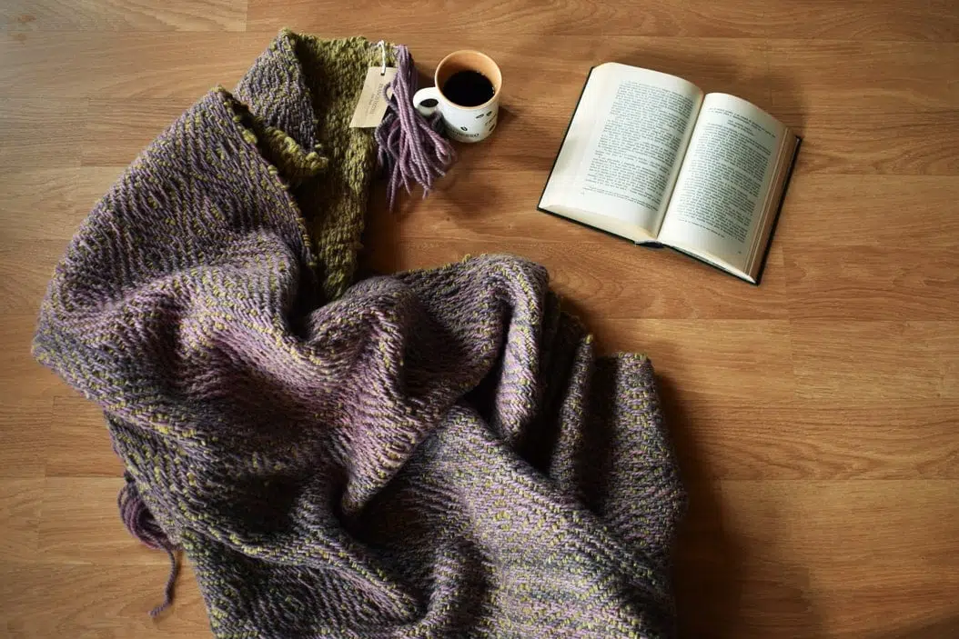 piecera de cama, Piecera tejida a telar piecera tejida, en color morado y un verde musgo hermoso, esta extendida en el suelo, asi puedes ver los dos diseños diferentes que posee este articulo ya que es reversible, junto a un libro y un cafe, para insinuar lo acogedor que resulta este articulo