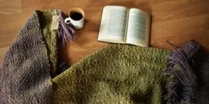 Piecera tejida a telar piecera tejida, en color morado y un verde musgo hermoso, esta extendida en el suelo, asi puedes ver los dos diseños diferentes que posee este articulo ya que es reversible, junto a un libro y un cafe, para insinuar lo acogedor que resulta este articulo