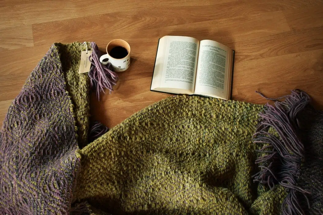 Piecera tejida a telar piecera tejida, en color morado y un verde musgo hermoso, esta extendida en el suelo, asi puedes ver los dos diseños diferentes que posee este articulo ya que es reversible, junto a un libro y un cafe, para insinuar lo acogedor que resulta este articulo