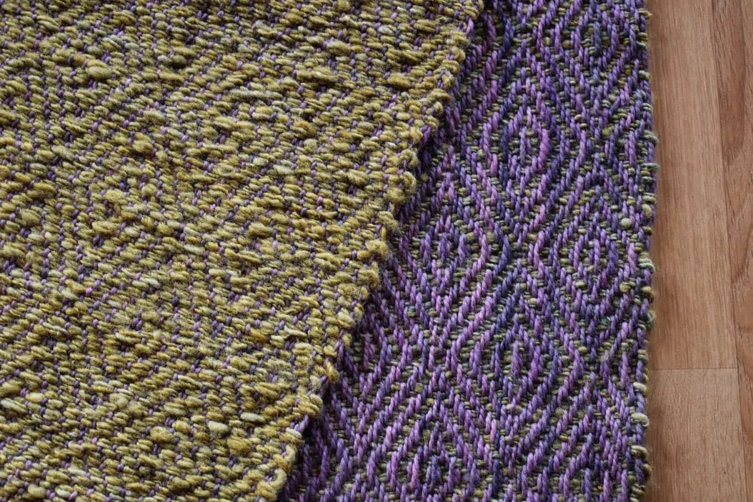 diseño de lana, piecera tejida, en color morado y un verde musgo hermoso, esta colgada sobre una baranda, asi puedes ver los dos diseños diferentes que posee este articulo ya que es reversible-