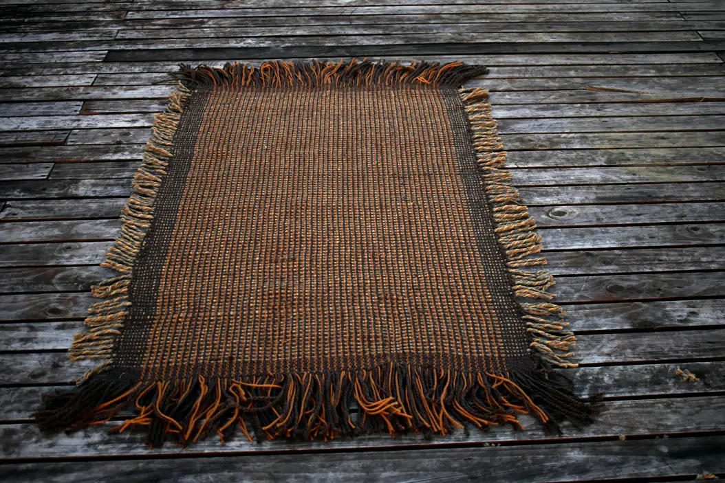 Vista completa de la alfombra artesanal de lana natural en colores cafés y grises, extendida sobre una tarima de madera